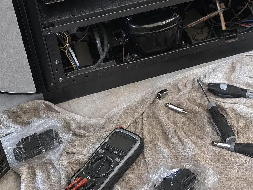 Bagged parts, a multimeter and screwdrivers on a towel in front of an open undercounter unit.