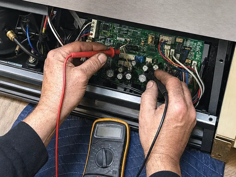 Technician's hands probing a green control board in the base of a stainless refrigerator, a yellow multimeter on a blue pad.