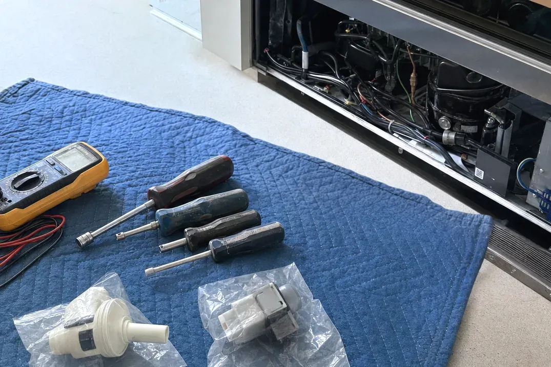 Multimeter, screwdrivers and bagged replacement parts on a blue pad beside an opened refrigerator base.